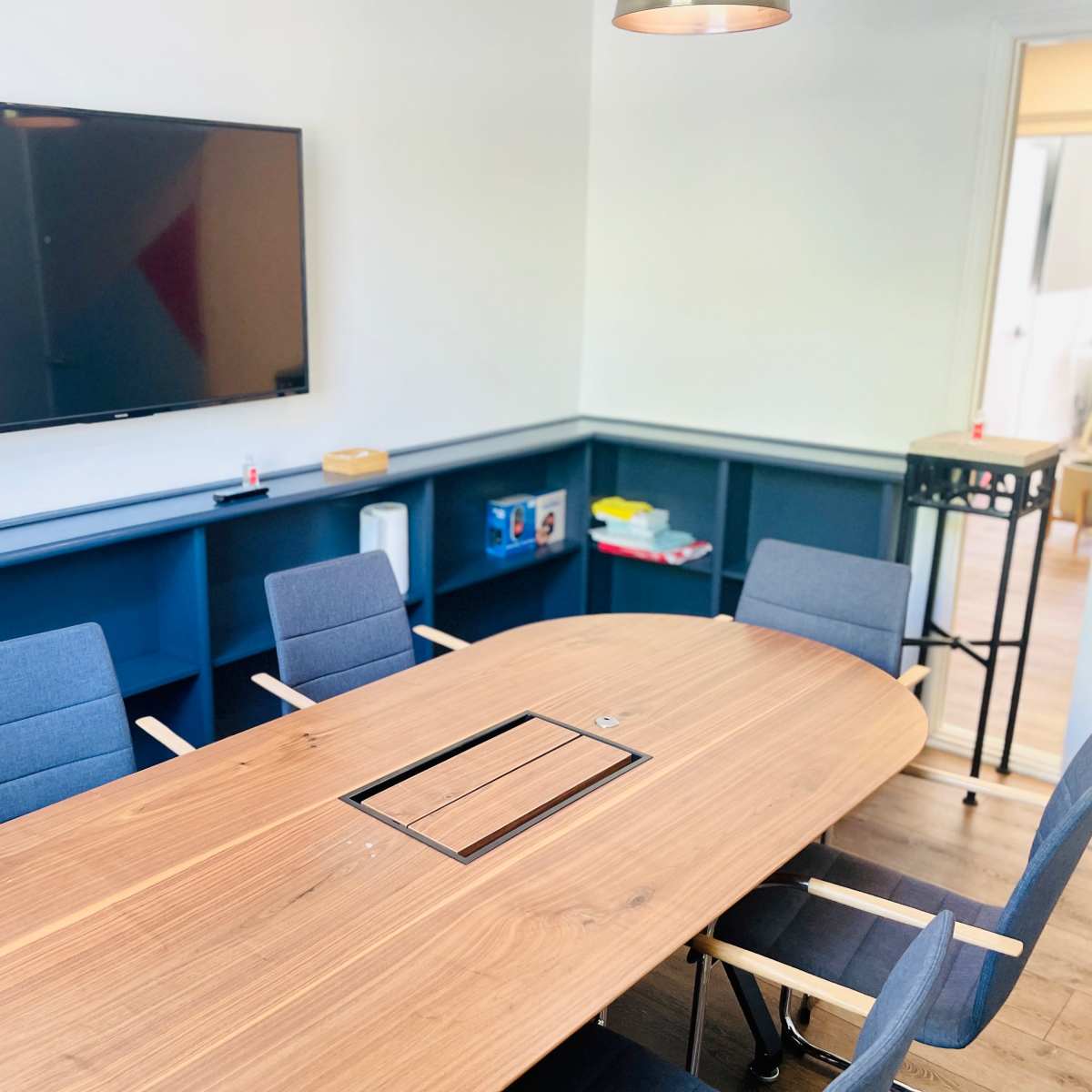 A modern conference room features a large wooden table surrounded by gray chairs and a wall-mounted television.