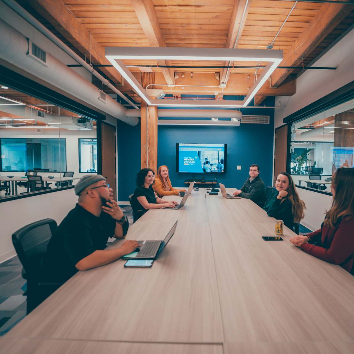 A group of six people sit around a long, wooden conference table in a modern office meeting room.