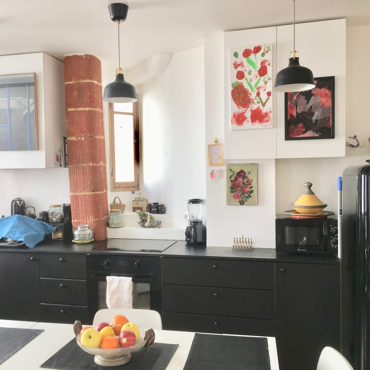 A modern kitchen features black cabinetry, white walls, and a central table with a fruit bowl, illuminated by pendant lamps.