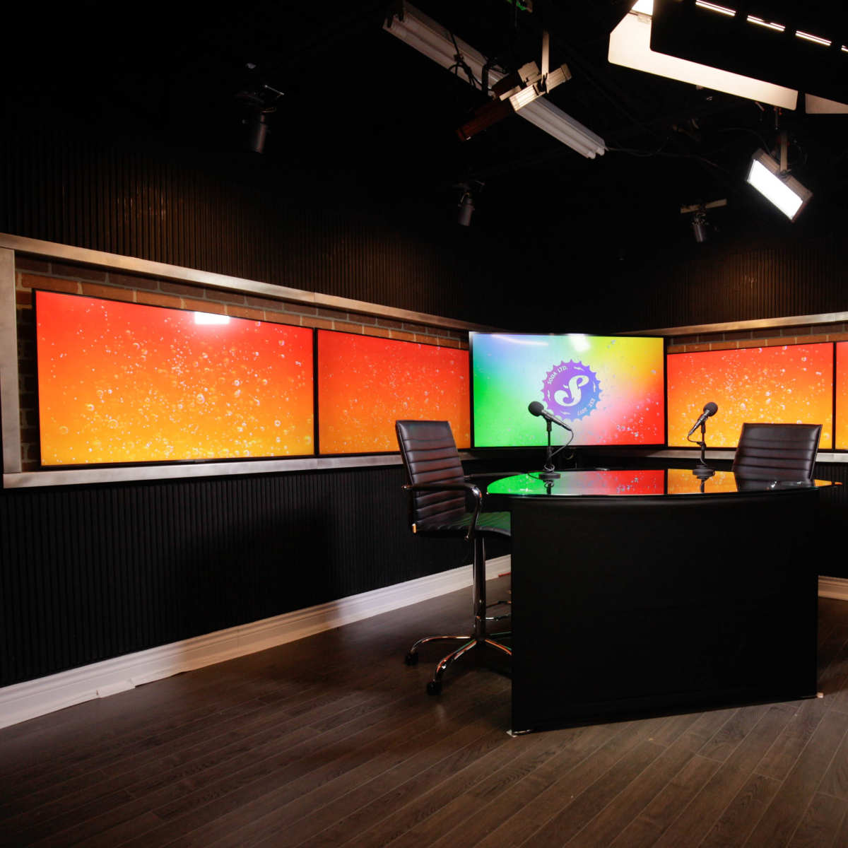 A modern studio setting features a central circular table with two chairs and multiple brightly colored screens in the background.