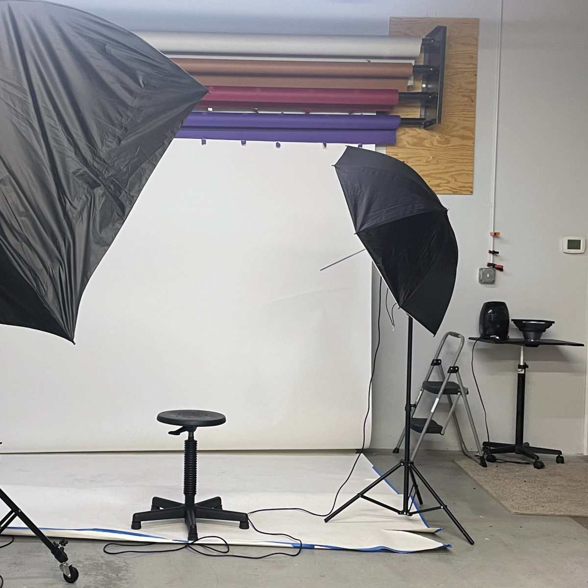 A photography studio setup with two umbrellas, a stool in the center, and various backgrounds hung on the wall.