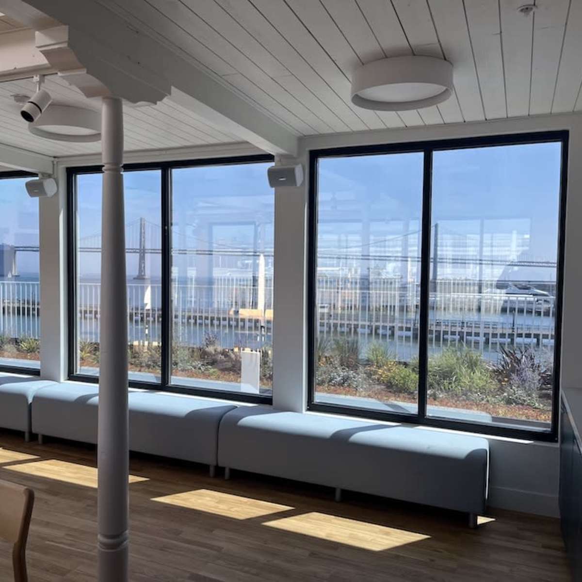 A bright interior space with large windows overlooking a body of water and a bridge in the background.