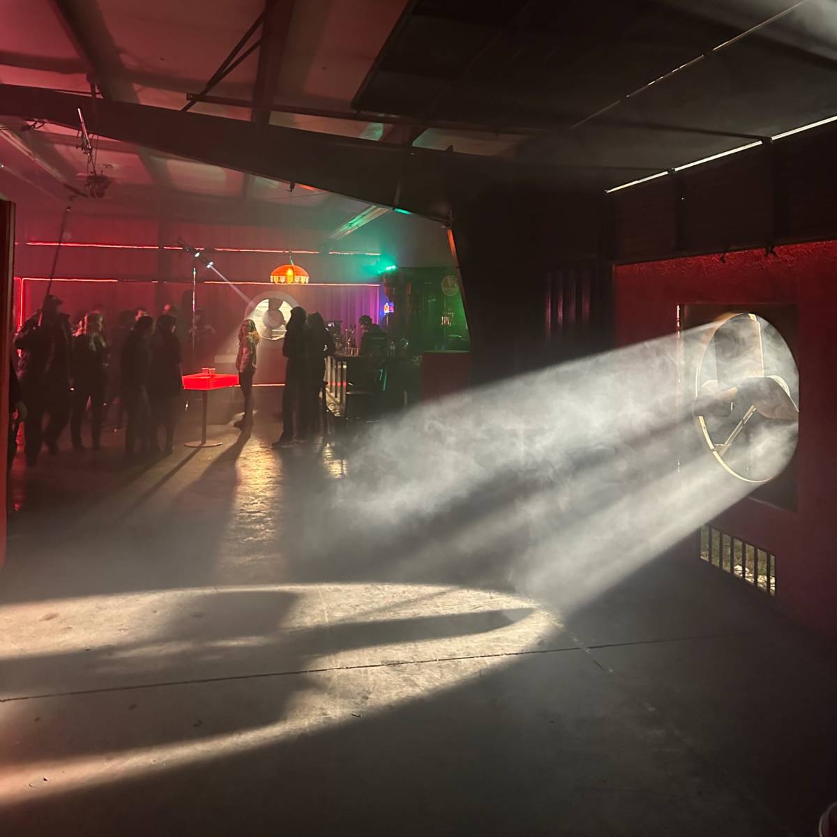 A dimly lit interior with smoke and beams of light streaming through a circular opening, illuminating a gathering of people in the background.