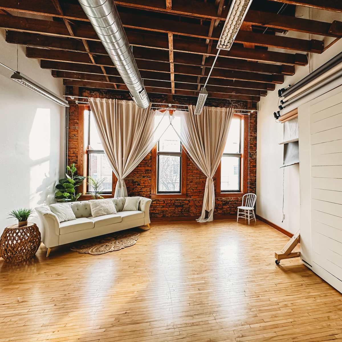 A spacious room with exposed brick walls, wooden flooring, large windows dressed with curtains, and minimal furniture including a white sofa and a chair.