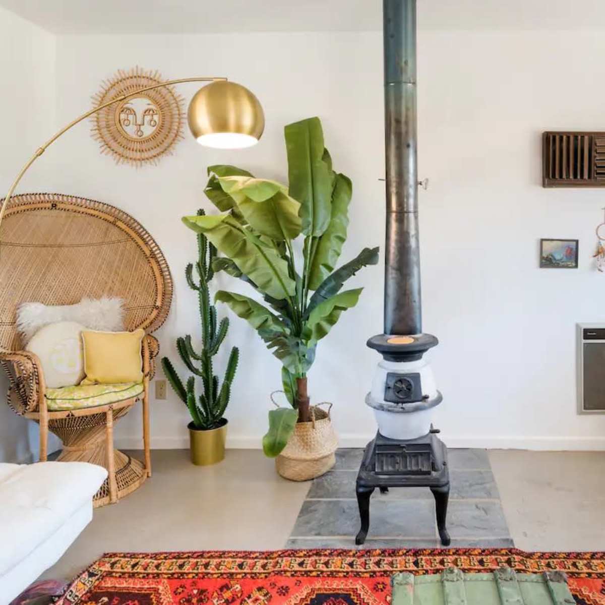 A cozy living room featuring a central wood stove, woven accent chair, decorative plants, and a combination of wall art.