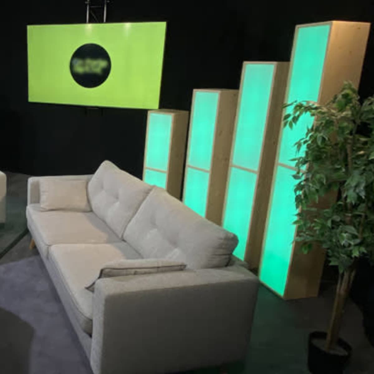 A minimalist lounge area with two gray sofas arranged around a light-colored wooden coffee table, set against a backdrop of illuminated green panels and a screen.