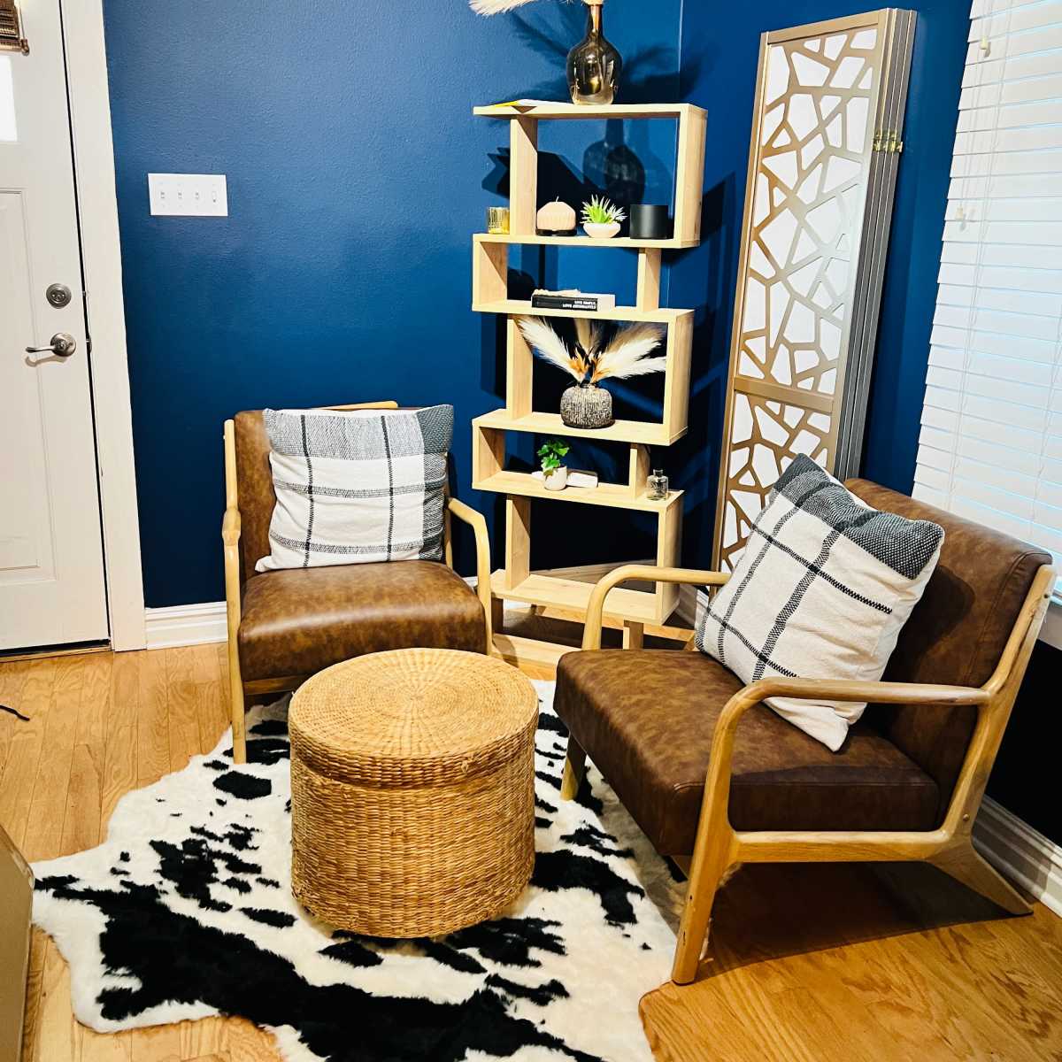 A cozy living space with two brown chairs, a round woven table, a black and white patterned rug, and a wooden shelf decorated with plants and a vase on a blue wall.
