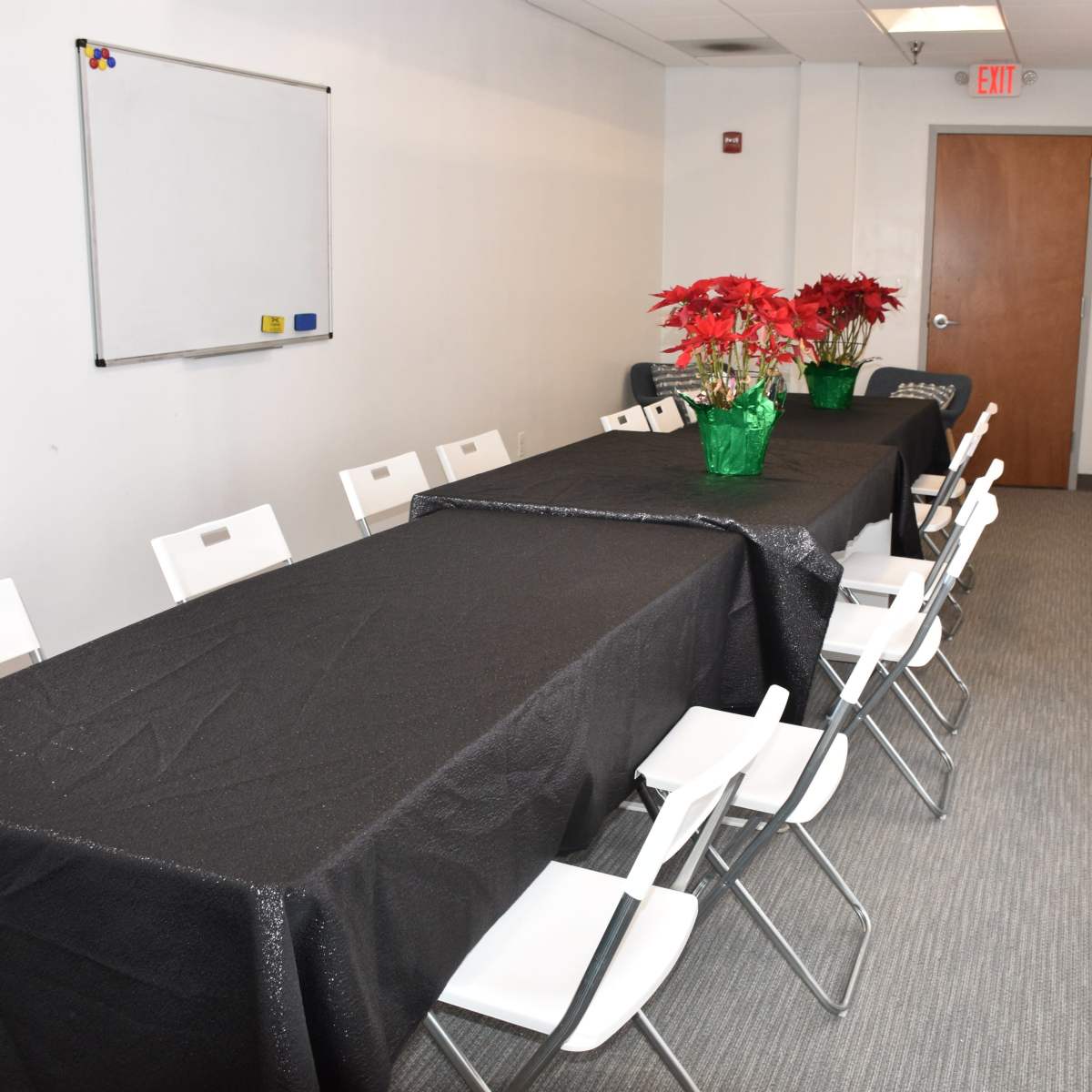 A long table covered with a black tablecloth is set in a conference room, lined with white chairs, and adorned with a centerpiece of potted red poinsettias.