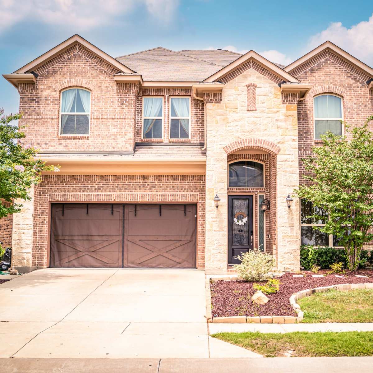 A two-story brick house with a stone facade and a front garage sits on a residential street, surrounded by manicured landscaping.
