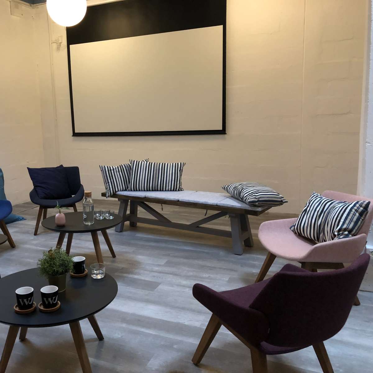 The scene shows a cozy lounge area with various chairs, a bench, tables, and a projector screen at the front.