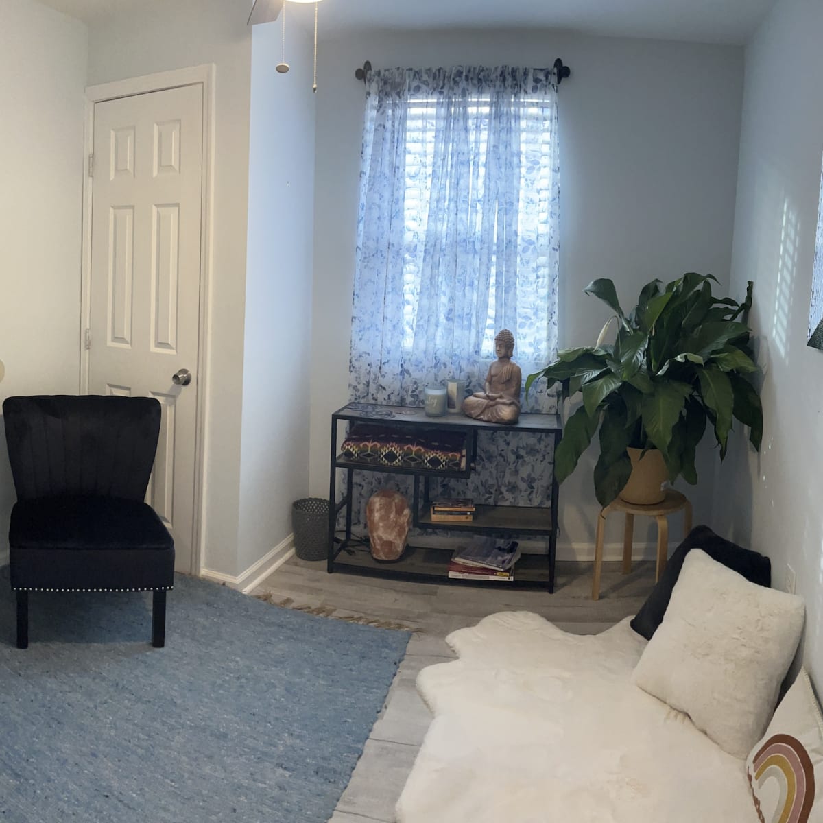 A small, well-lit room features two black chairs, a white area rug, and a bookcase with decorative items, including a statue of a Buddha and a large plant near a window.