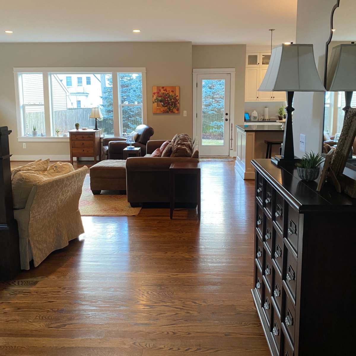 A living room area with large windows, hardwood flooring, and furniture including a sofa, chairs, and a wooden cabinet.