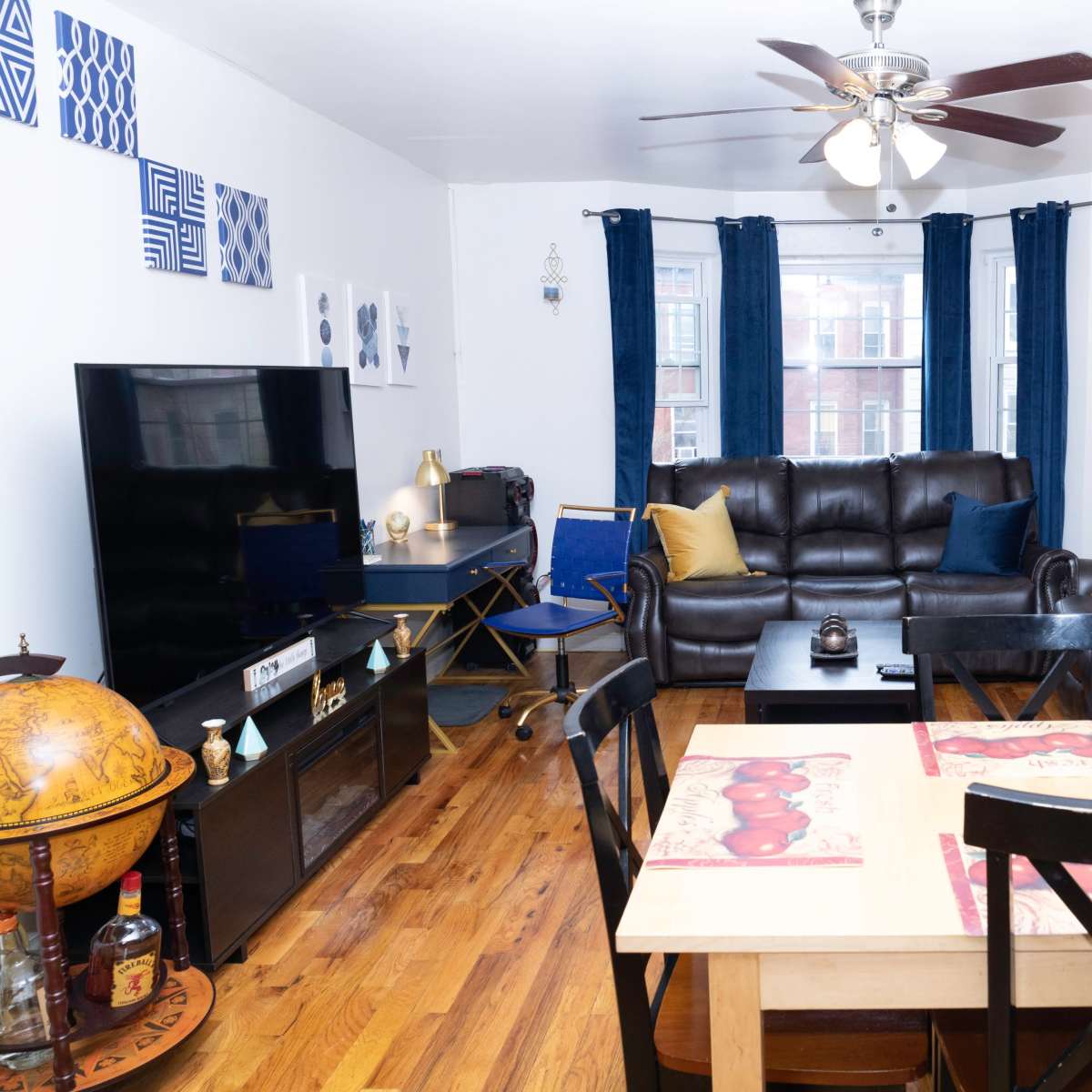 The living room features two brown leather sofas, a black television stand, a dining table with black chairs, and decor including blue curtains and a world map on the wall.