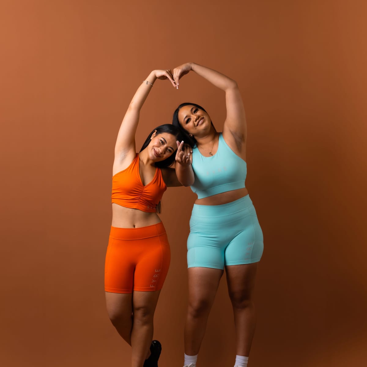 Two women in brightly colored athletic wear make a heart shape with their arms against a brown background.