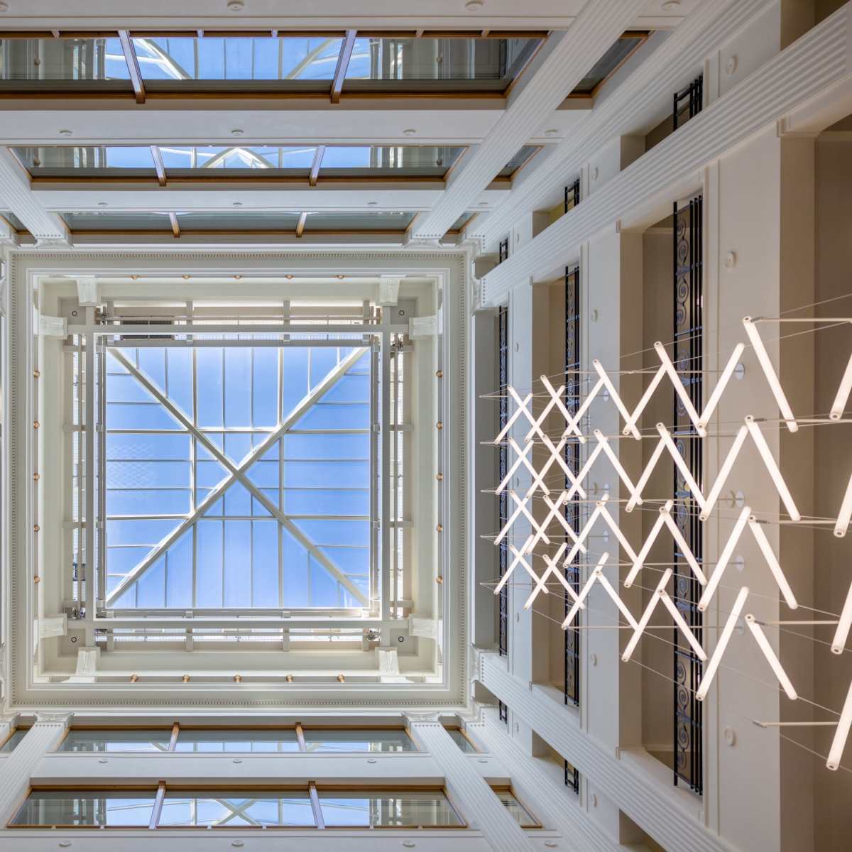 An interior atrium with a glass ceiling and linear lighting, viewed from below.