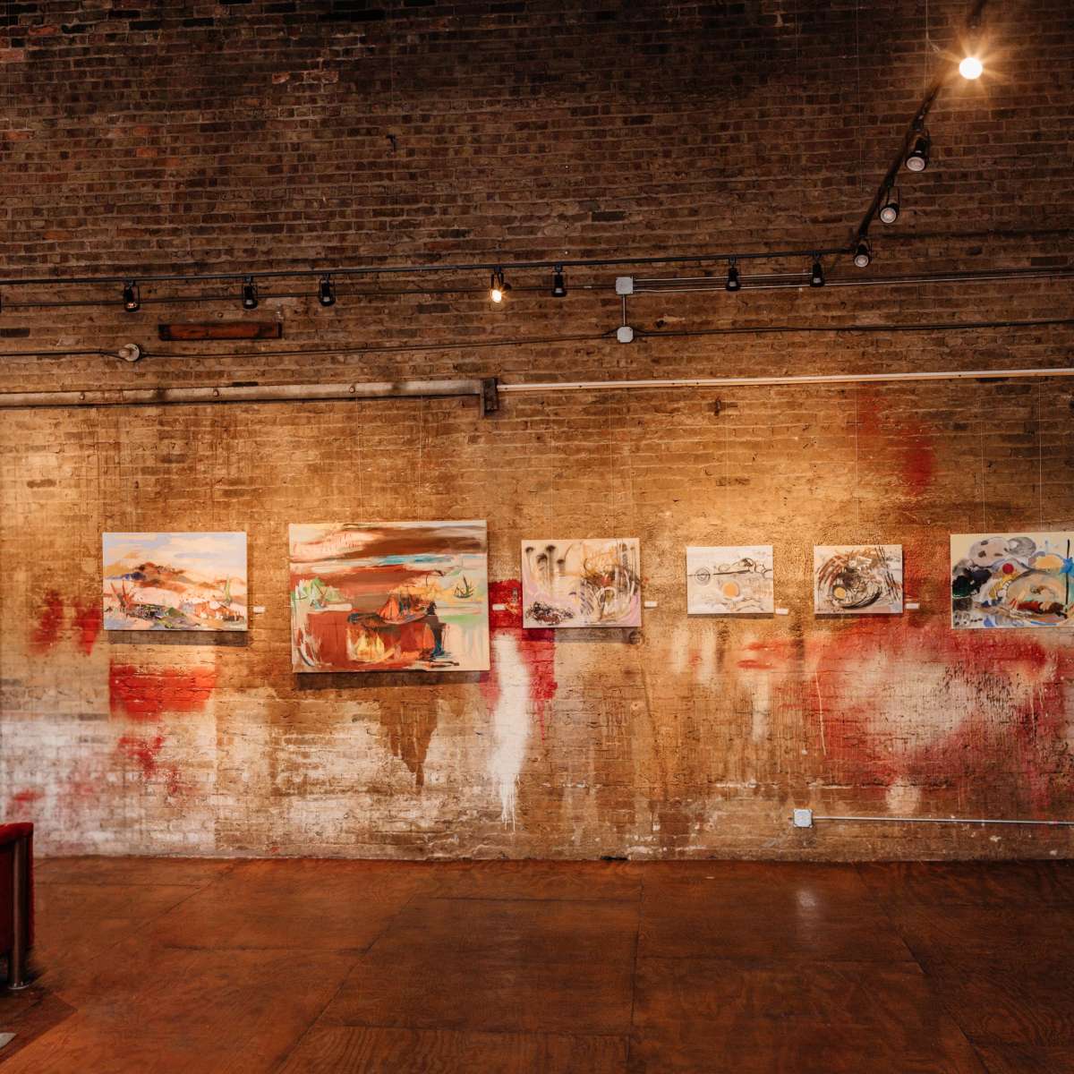 A gallery interior with a brick wall displaying several abstract paintings and a red velvet couch in the foreground.