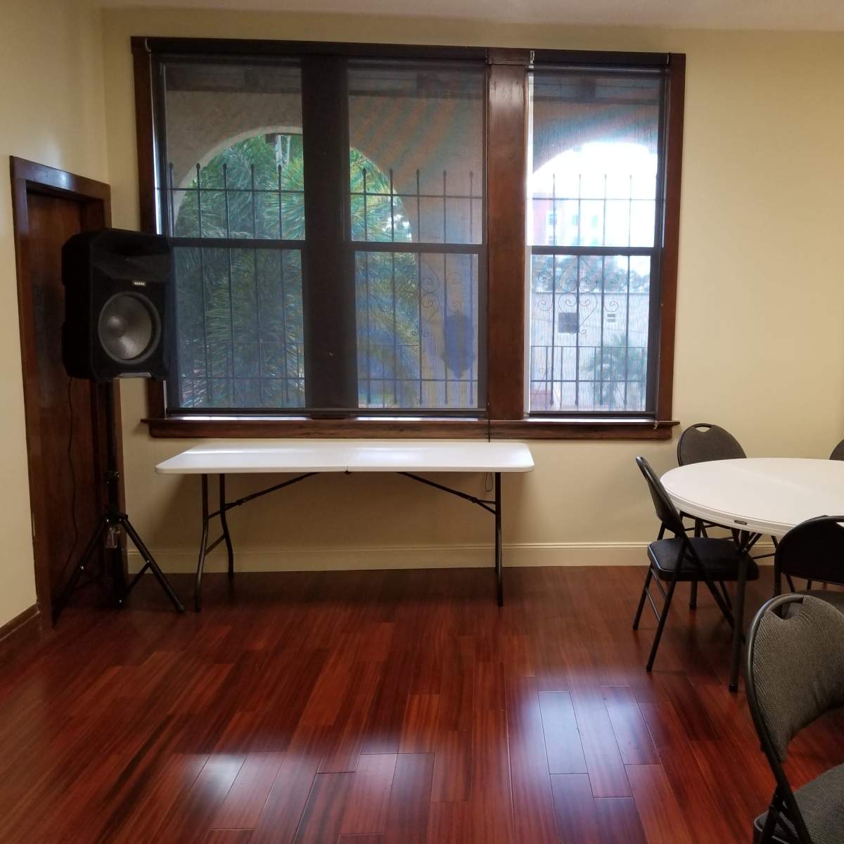 A room with wooden flooring, two round tables with chairs, a folding table, and a speaker next to the windows.