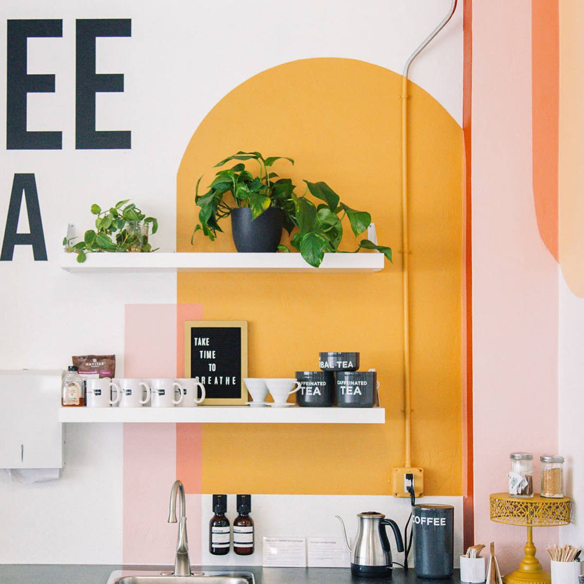A café interior with a wall featuring bold text that reads "COFFEE AND TEA" alongside shelves with plants and various coffee-related items.