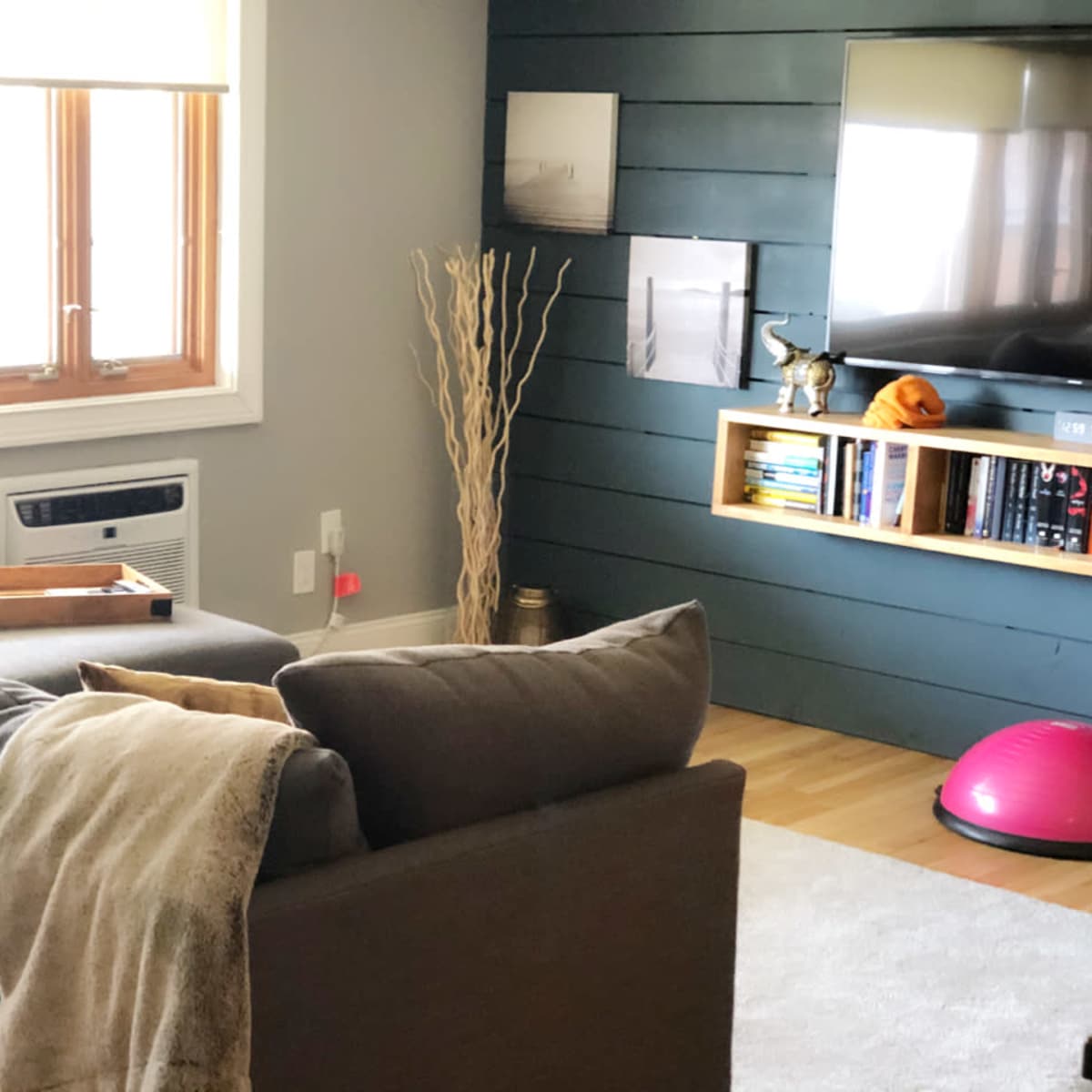 A living room features a gray sofa with cushions, an air conditioning unit, a television mounted on a dark wall, and shelves with books and decorative items.