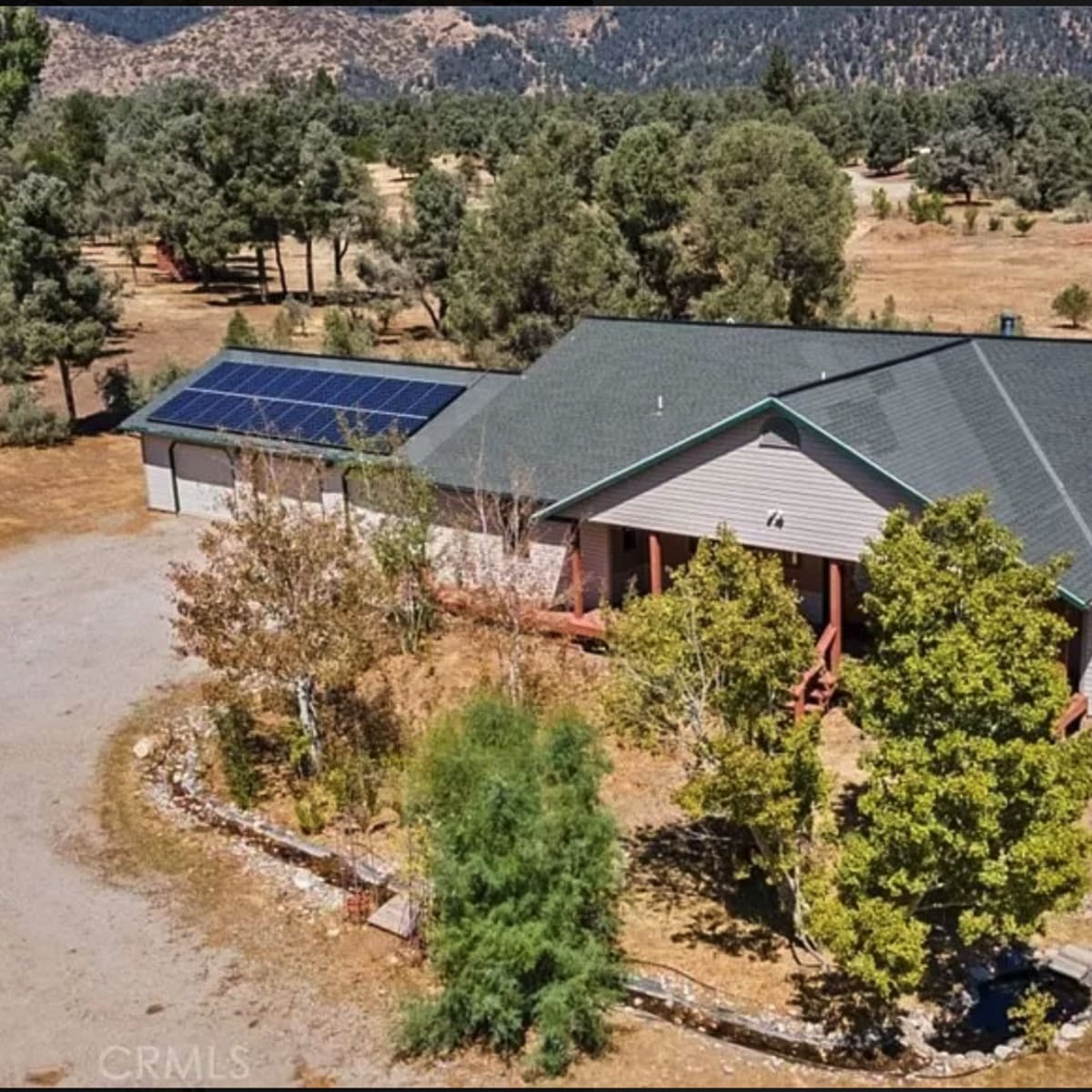 A single-story house with a solar panel on the roof, surrounded by trees and a large open area.