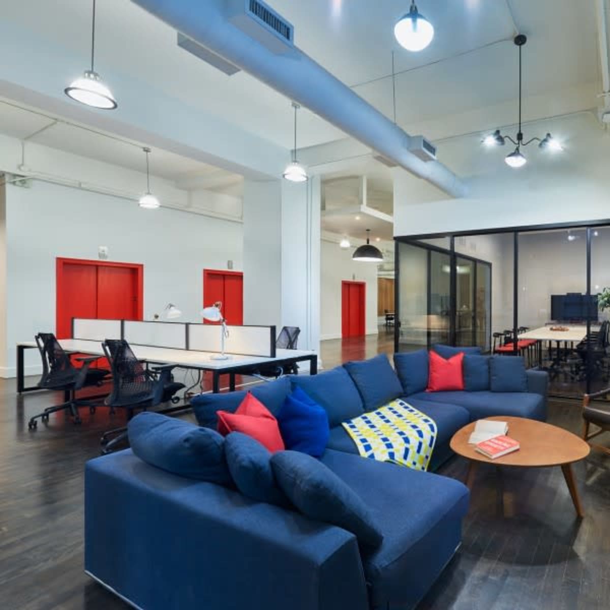 A modern office space featuring a large blue sectional sofa, a coffee table, and workstations with black office chairs, alongside large windows and red accent doors.