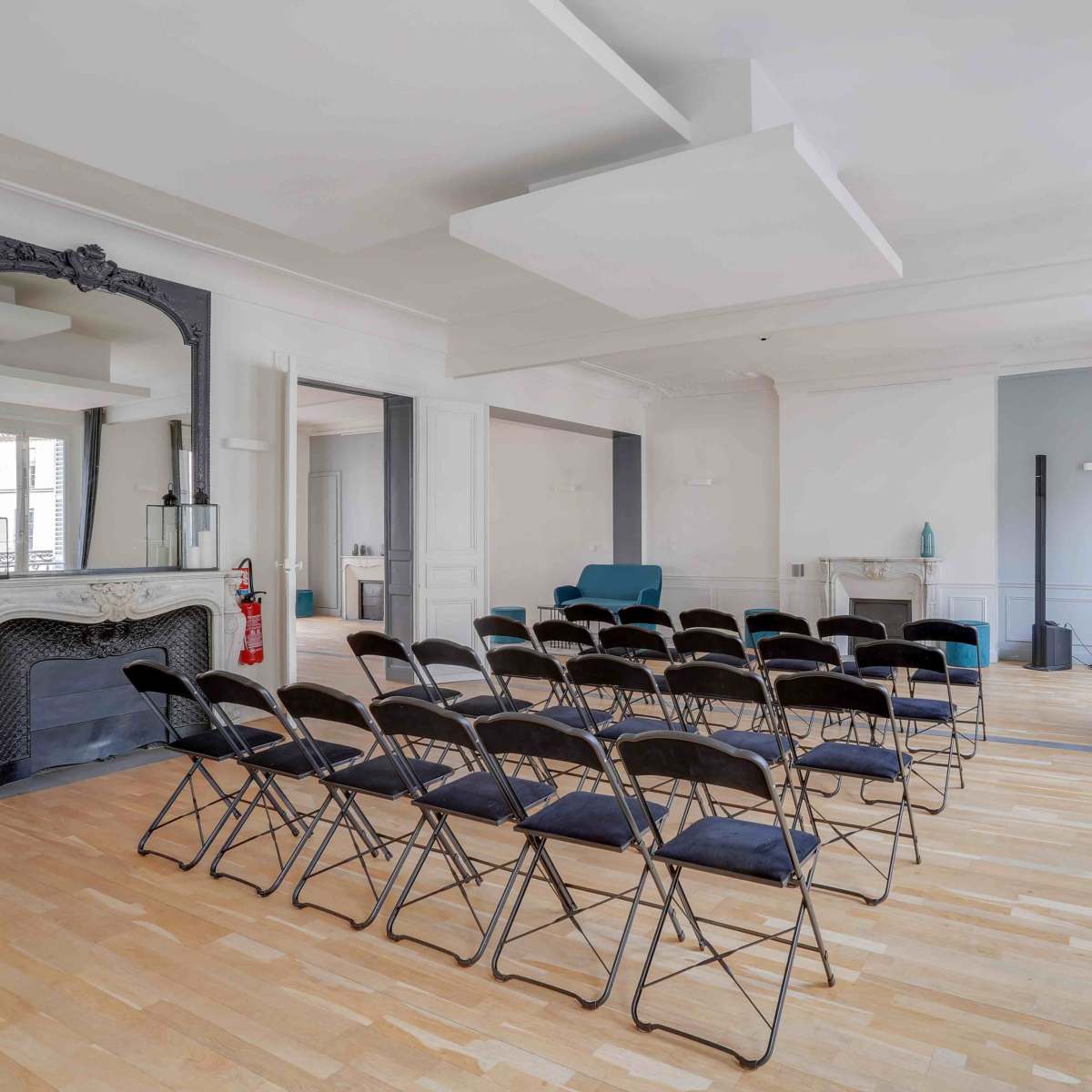 A spacious room with a row of black folding chairs arranged facing a light blue wall, with a fireplace and large windows dressed in gray curtains.