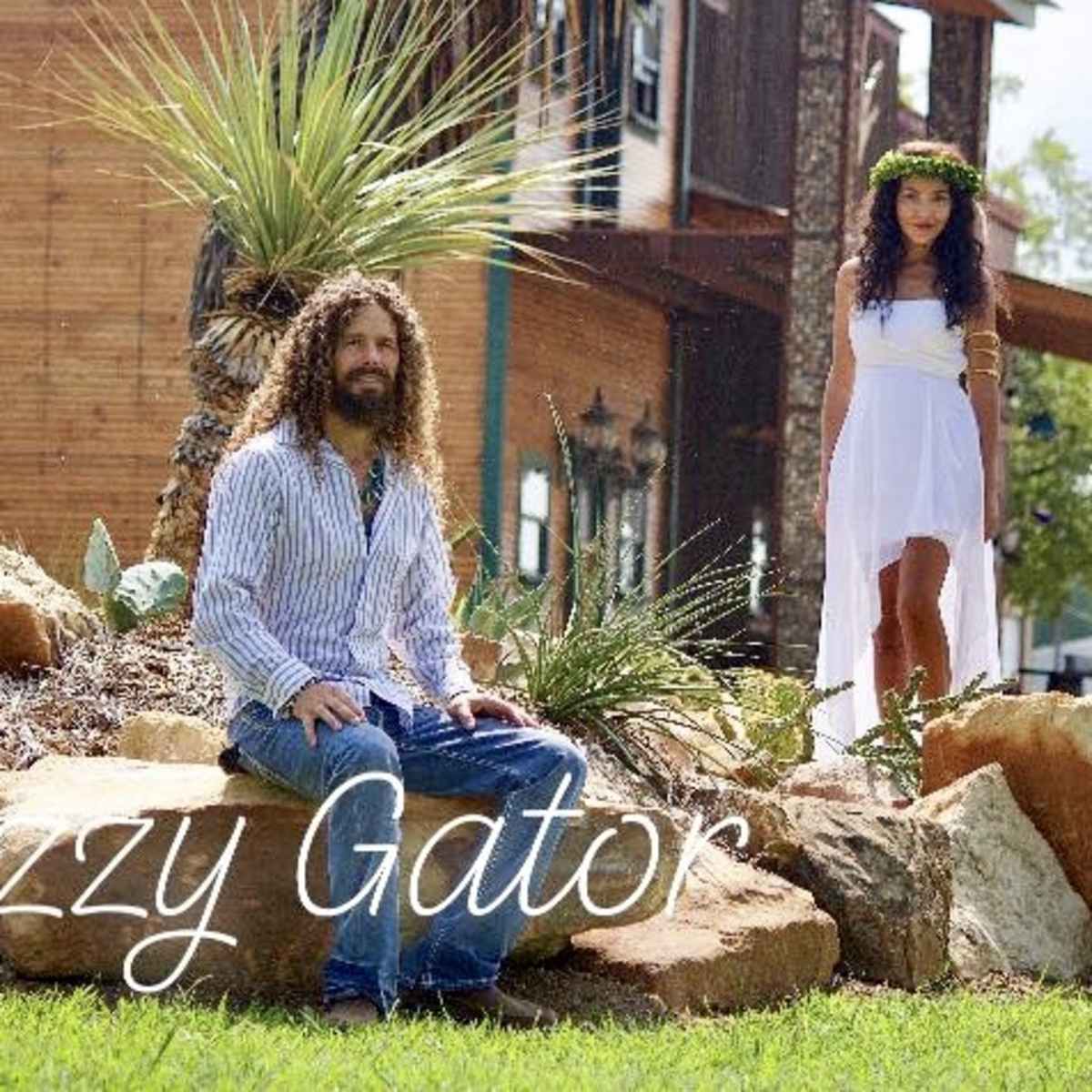 A man with long hair sits on a rock while a woman in a white dress stands nearby in a landscaped yard with plants and a wooden building in the background.