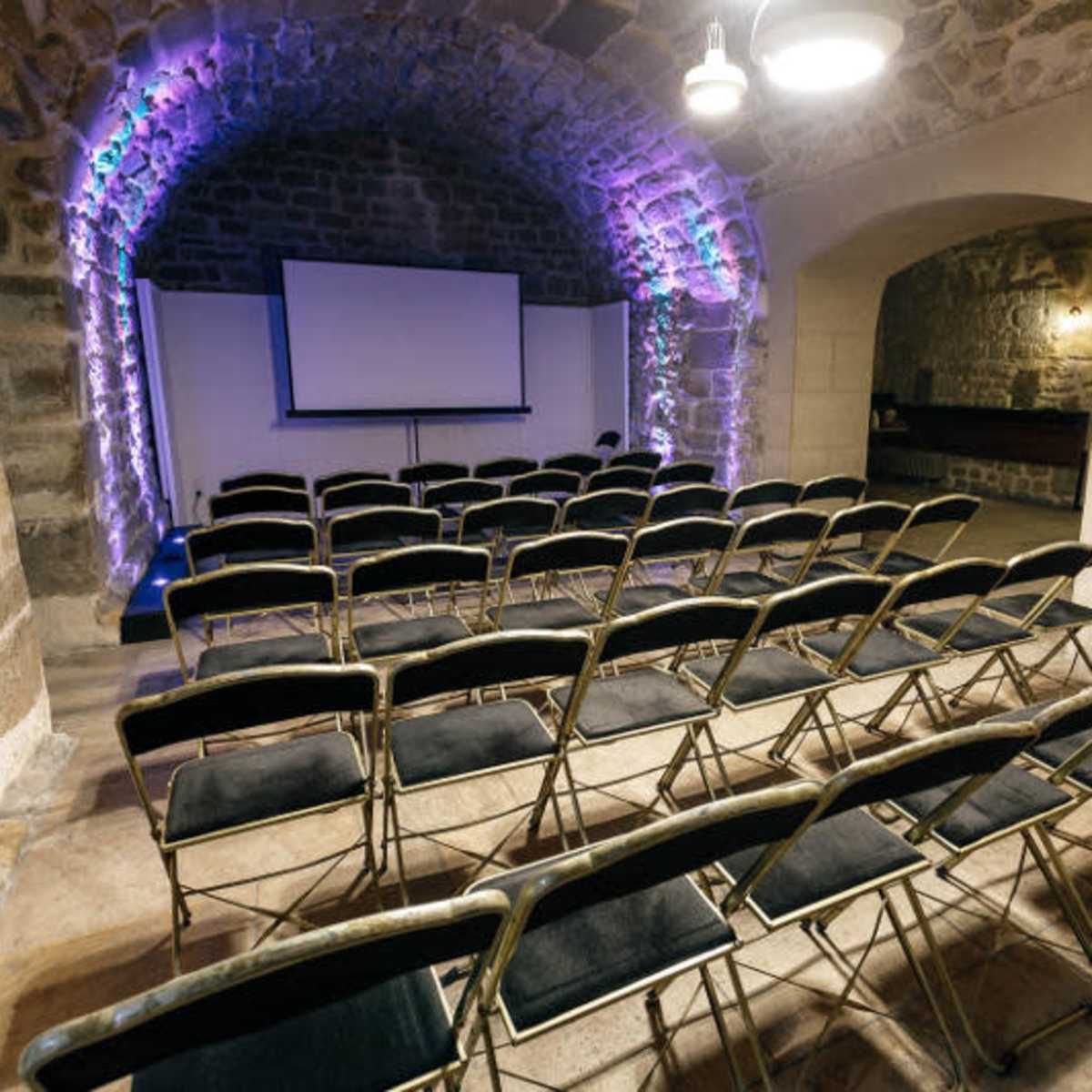A stone-walled meeting room features rows of black folding chairs facing a projection screen.
