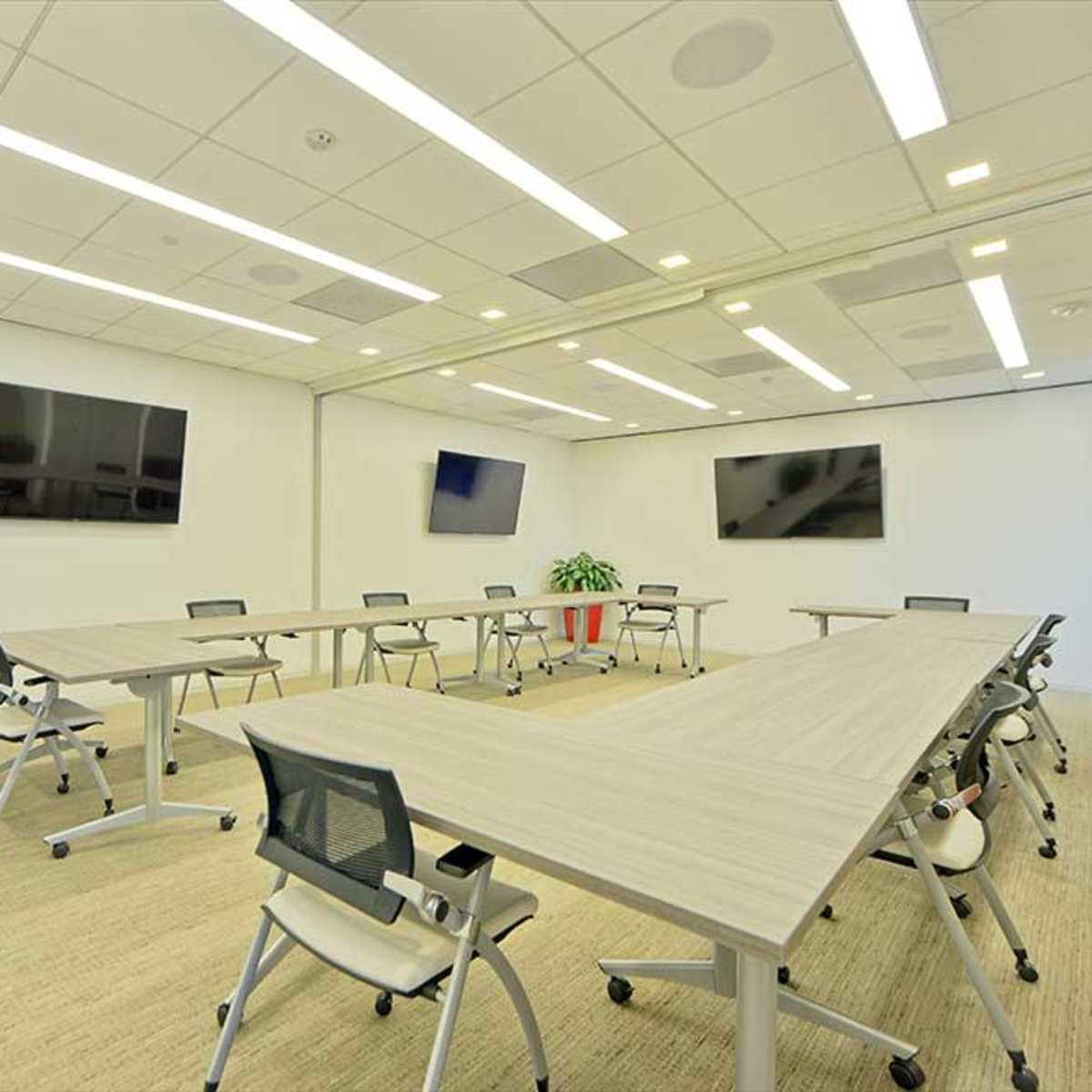 A modern conference room with multiple tables arranged in a U-shape, several televisions mounted on the walls, and a large potted plant in the corner.