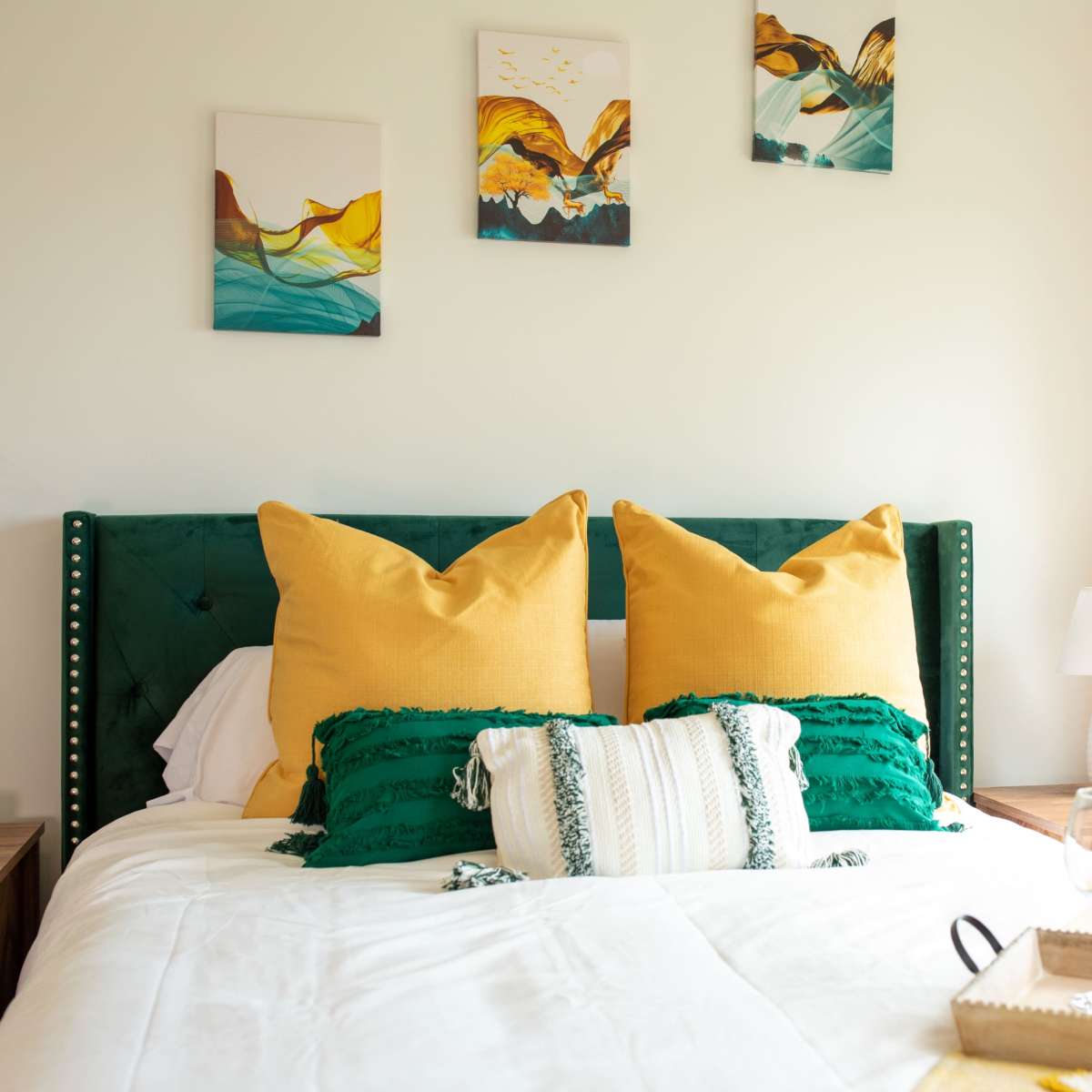 A neatly made bed features vibrant yellow pillows and a decorative throw, with a tray holding a bottle of wine and glasses set beside a window with yellow curtains.