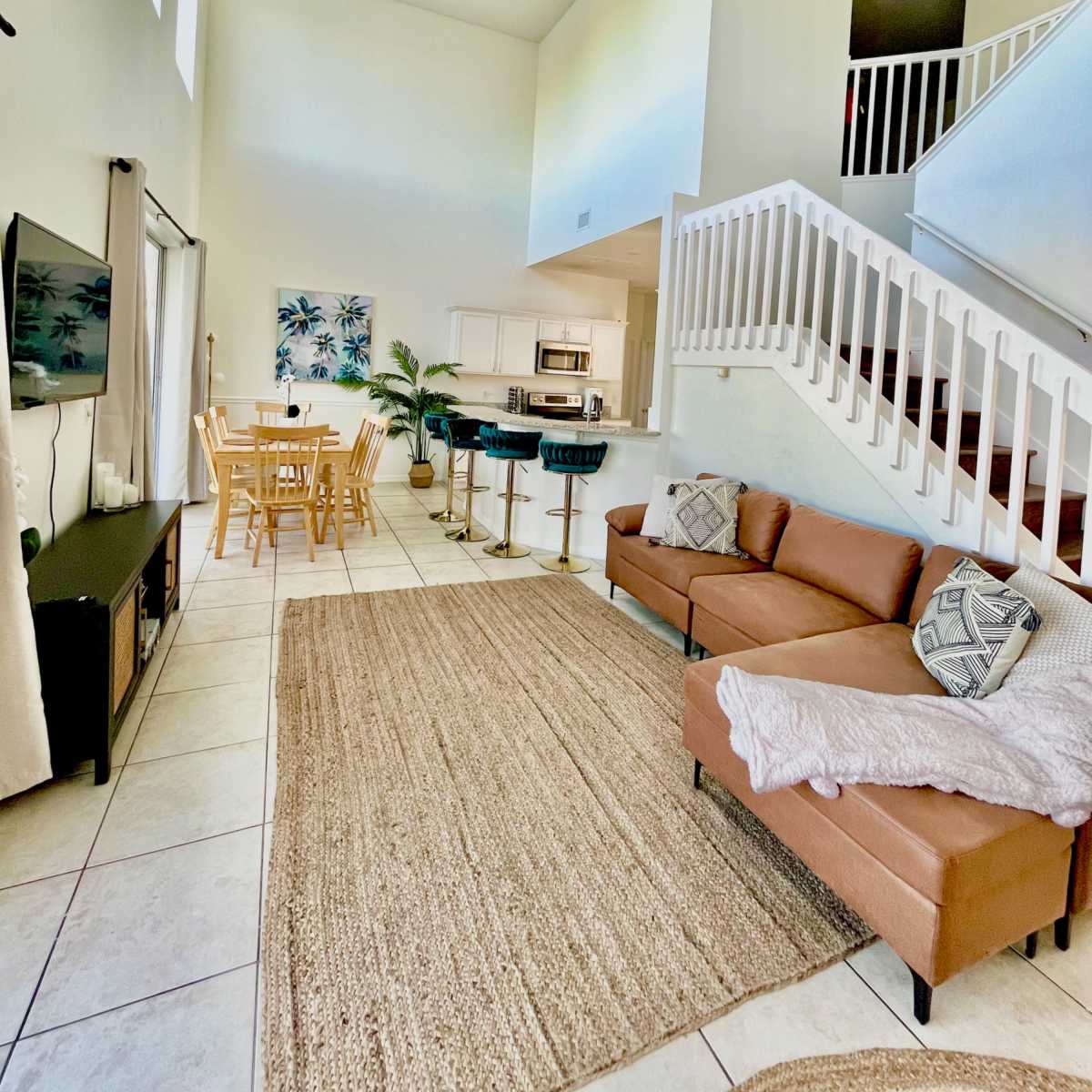 A brightly lit open living space with a staircase, a seating area with a brown couch, a dining table for six, and a kitchen visible in the background.