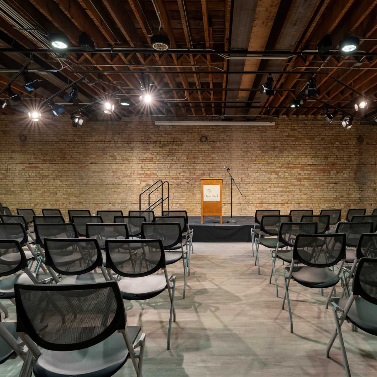 A spacious indoor auditorium with a podium and microphone at the front, facing a set of empty black chairs arranged in rows.