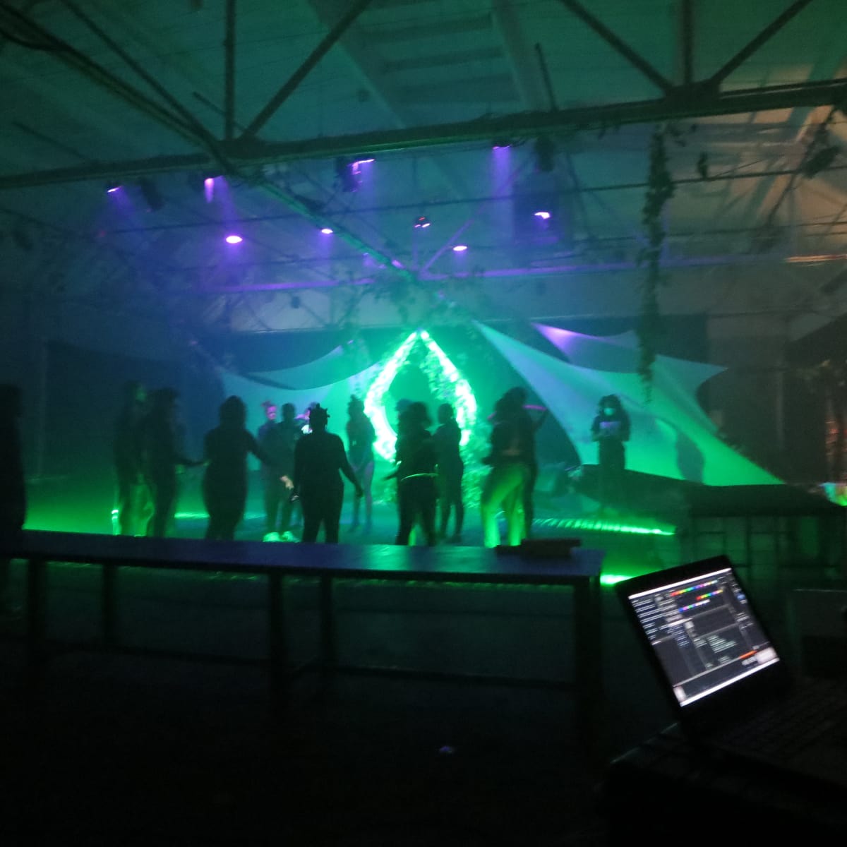 A group of people dances under purple and green lights in an industrial space.