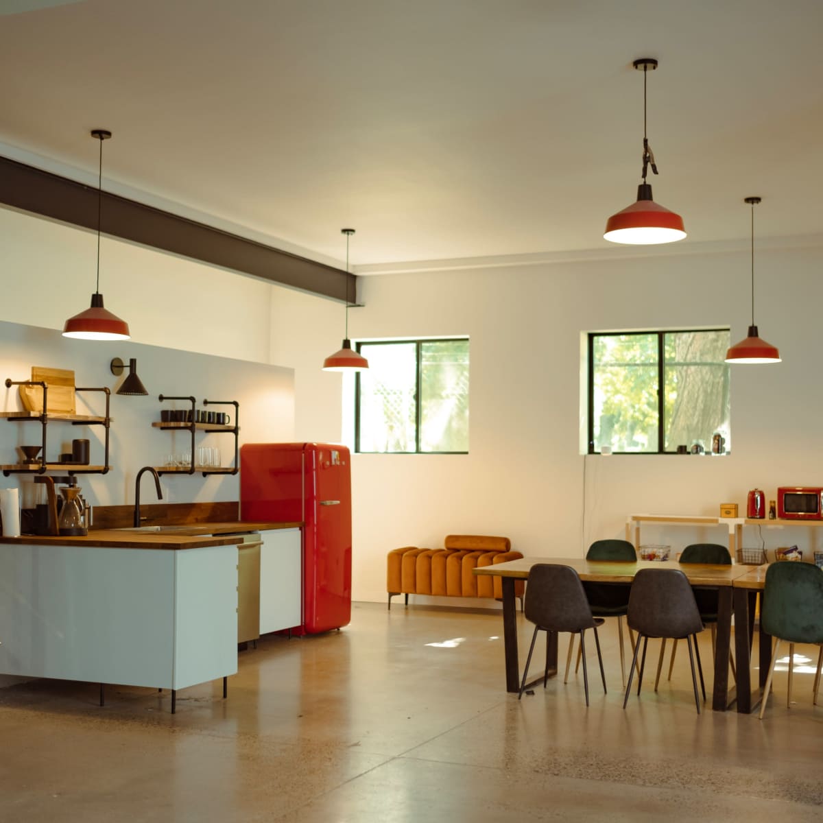 A spacious modern kitchen and dining area with a red refrigerator, wooden dining table, and large windows allowing natural light to enter.