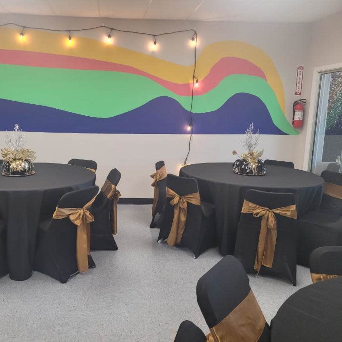 A decorated indoor event space with two round tables covered in black tablecloths and gold bows on black chairs, set against a colorful mural and string lights.
