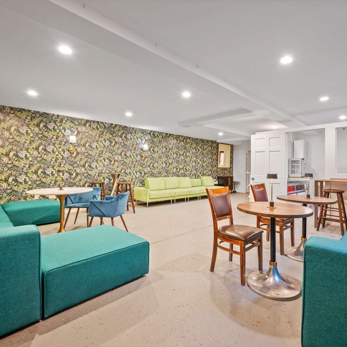 A brightly lit communal room with teal sofas, wooden tables, and a patterned wall, featuring a mix of seating arrangements.