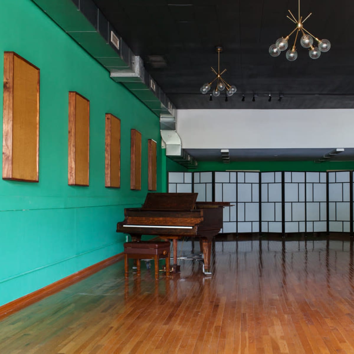 An empty room with polished wooden floors, a green accent wall with wooden panels, and a grand piano near the center, illuminated by modern light fixtures.
