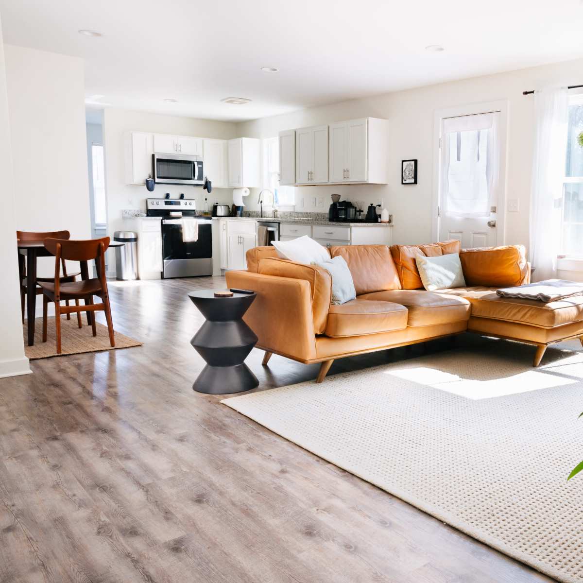 A bright, modern living room with a tan leather sofa, a round side table, a woven area rug, and plants, leading into a minimalist kitchen area.