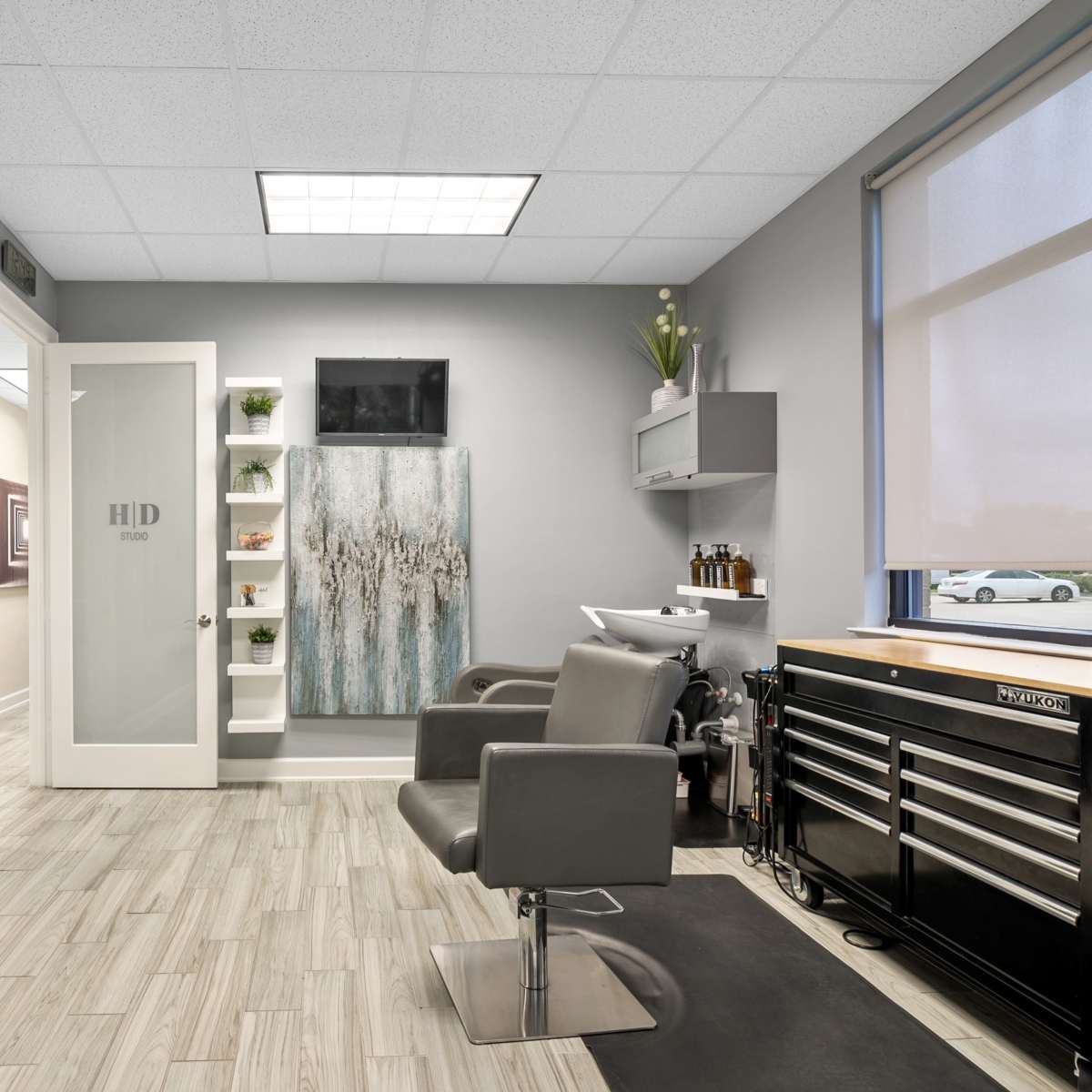 A modern hair salon with a gray and white color scheme, featuring two styling chairs, a large mirror, shelf storage, and a door labeled "HD" leading to another area.