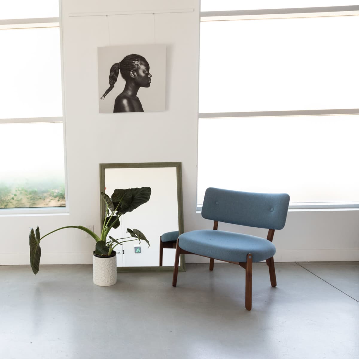 A blue chair is positioned beside a potted plant and a mirror in a brightly lit room with large windows.