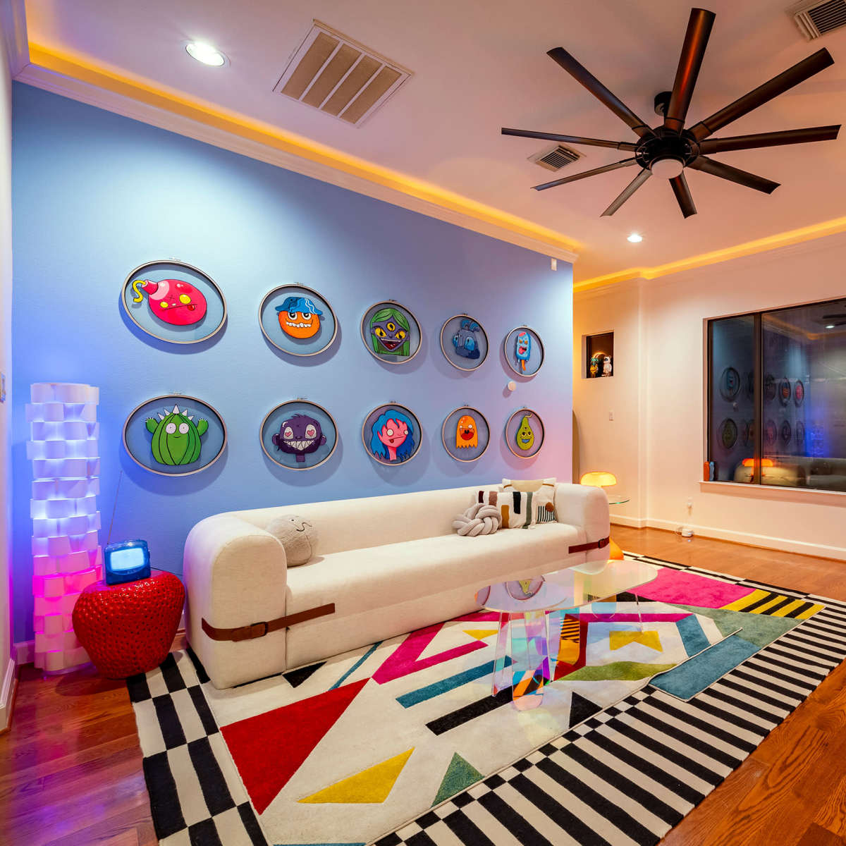 The room features a modern white sofa, colorful wall art, a circular red chair, and a patterned rug on hardwood flooring.