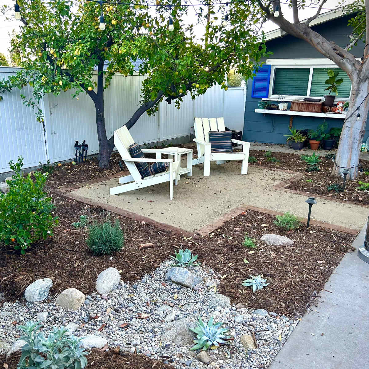 A landscaped backyard features two white chairs on a sandy area surrounded by gravel, rocks, and various plants, with a tree and a house in the background.