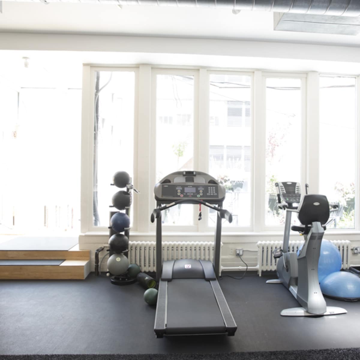 A bright gym interior with a treadmill, stationary bike, and exercise balls against large windows.