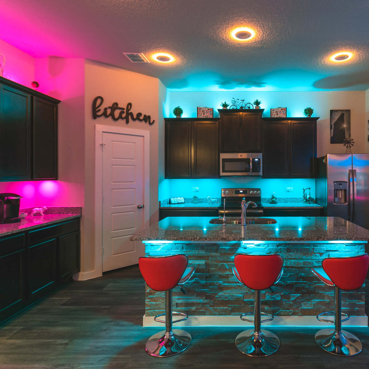 A modern kitchen features dark cabinetry, a granite countertop, and illuminated shelving with blue and pink lighting.
