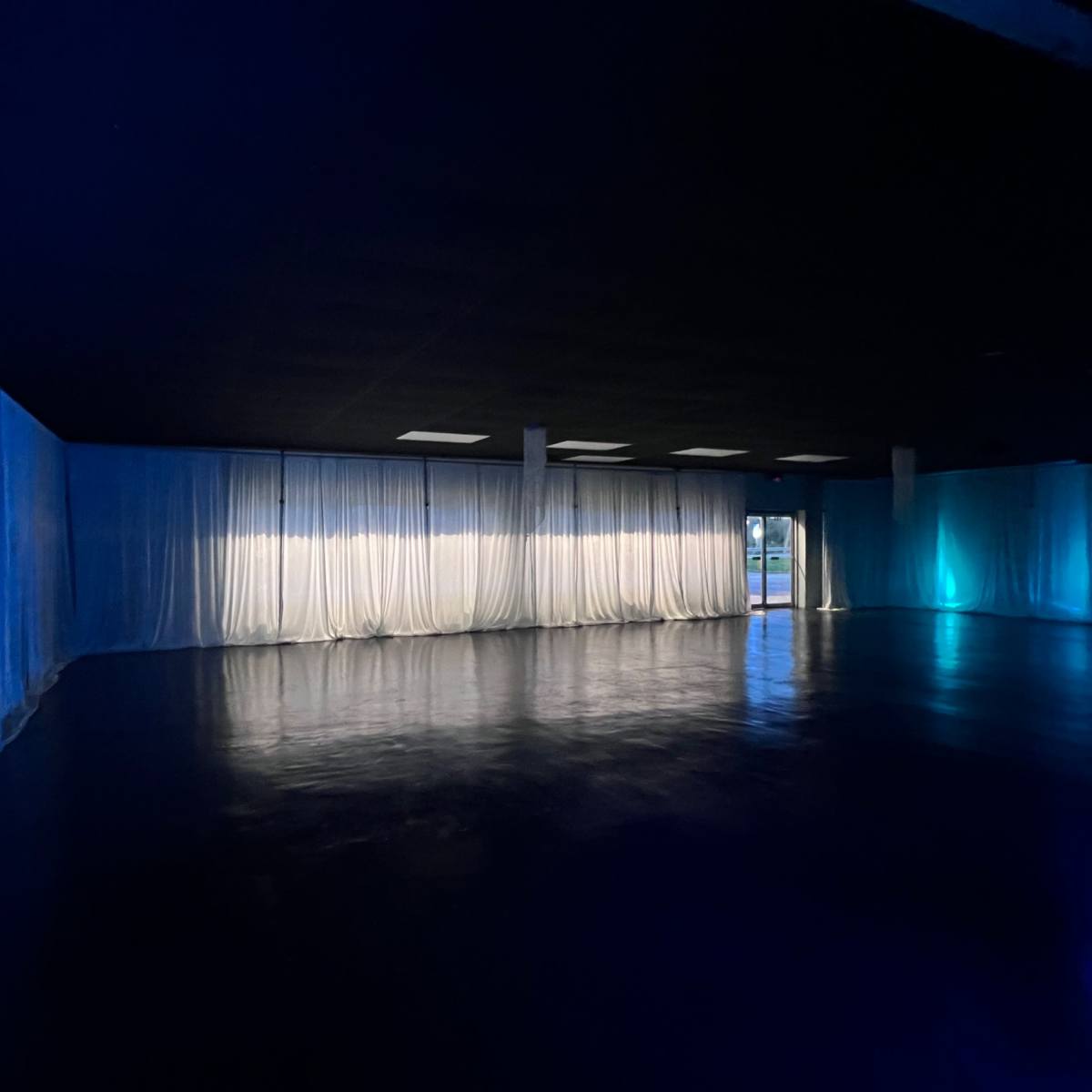 An empty room with dark flooring and blue-lit sheer curtains lining the walls.