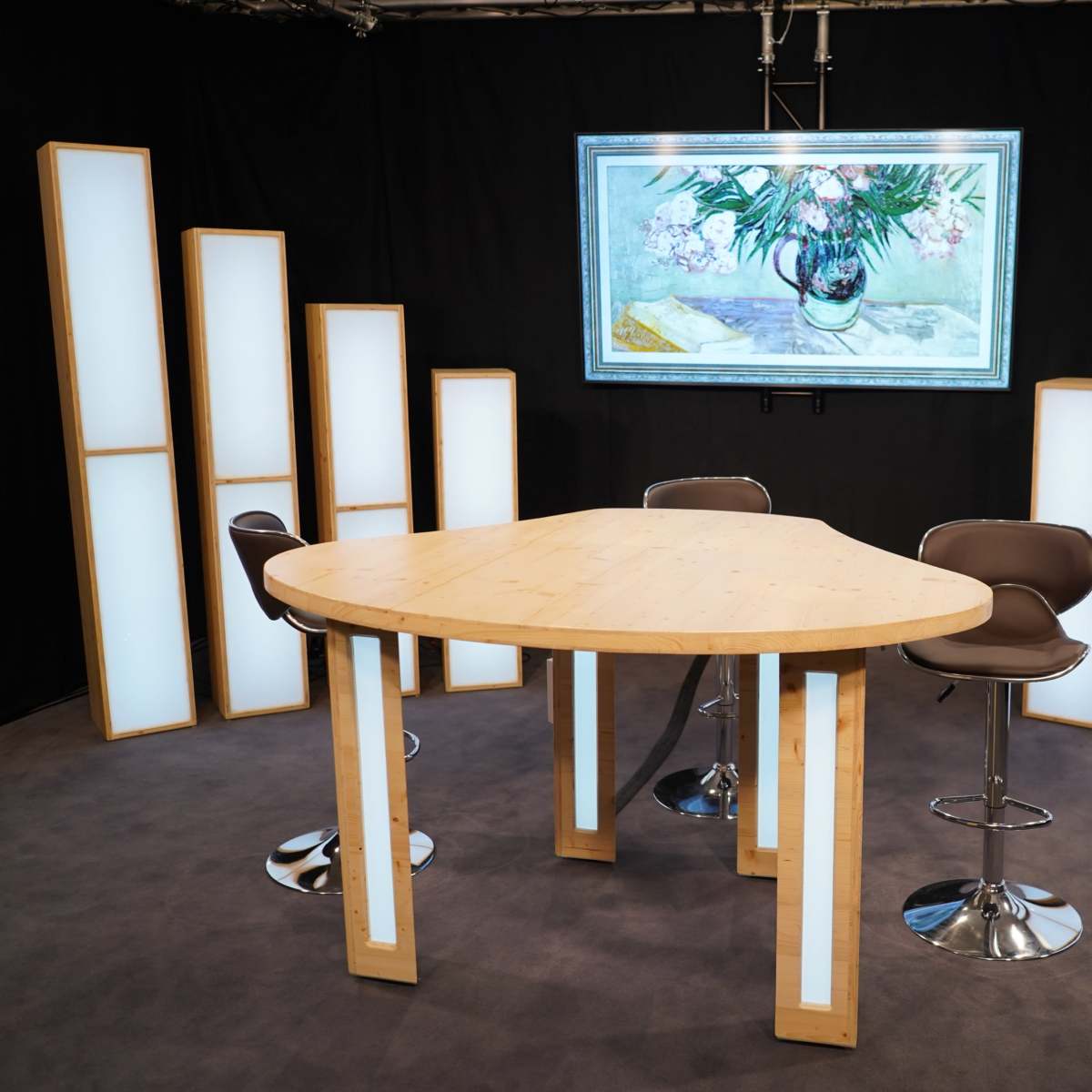 A modern studio setup features a triangular wooden table surrounded by high stools, illuminated by tall, backlit panels and a large artwork displayed above.