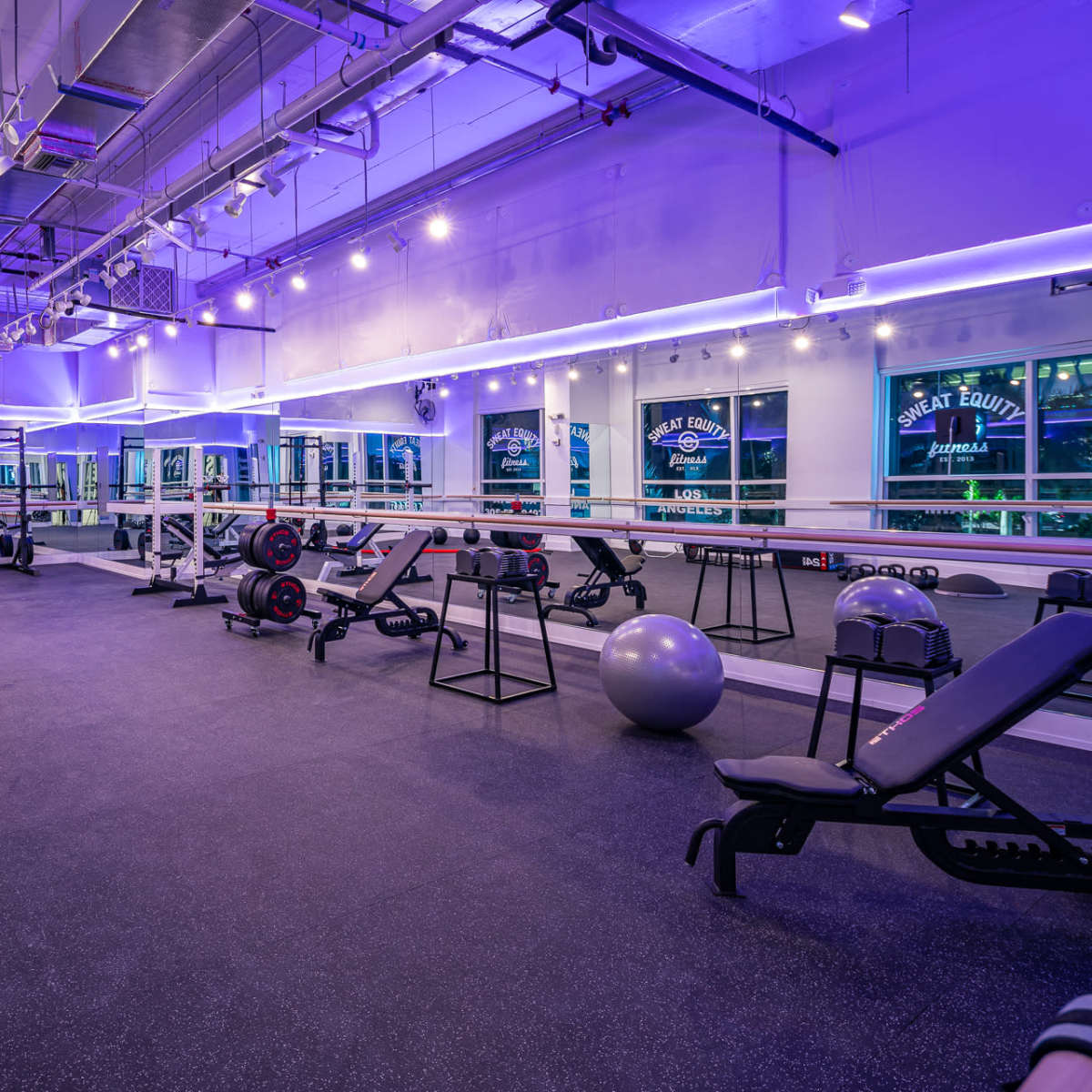 A well-equipped fitness studio featuring exercise machines, free weights, and various fitness accessories, illuminated by purple lights.