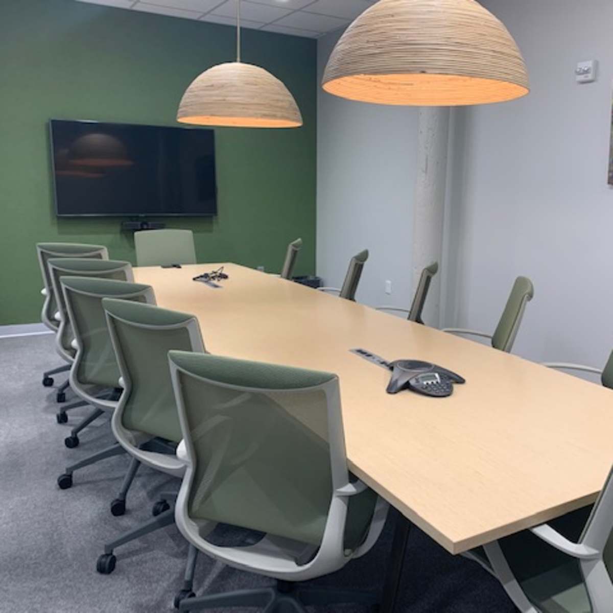 A long wooden conference table with eight green office chairs is positioned under two pendant lights in a modern meeting room adorned with a green accent wall.