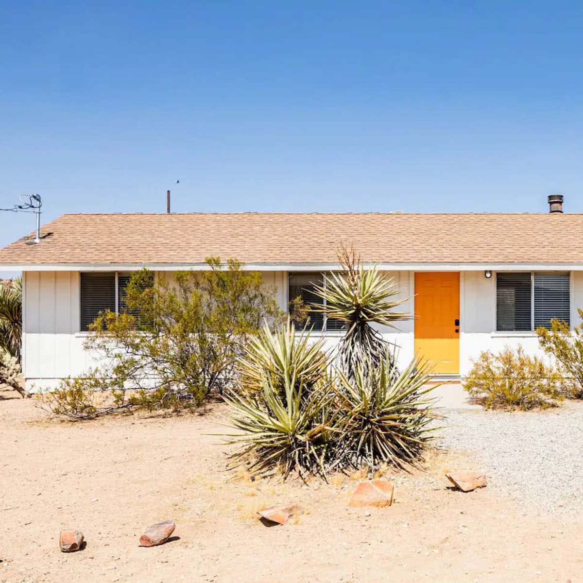A single-story, light-colored house with a bright orange front door is surrounded by desert vegetation and mountains under a clear blue sky.