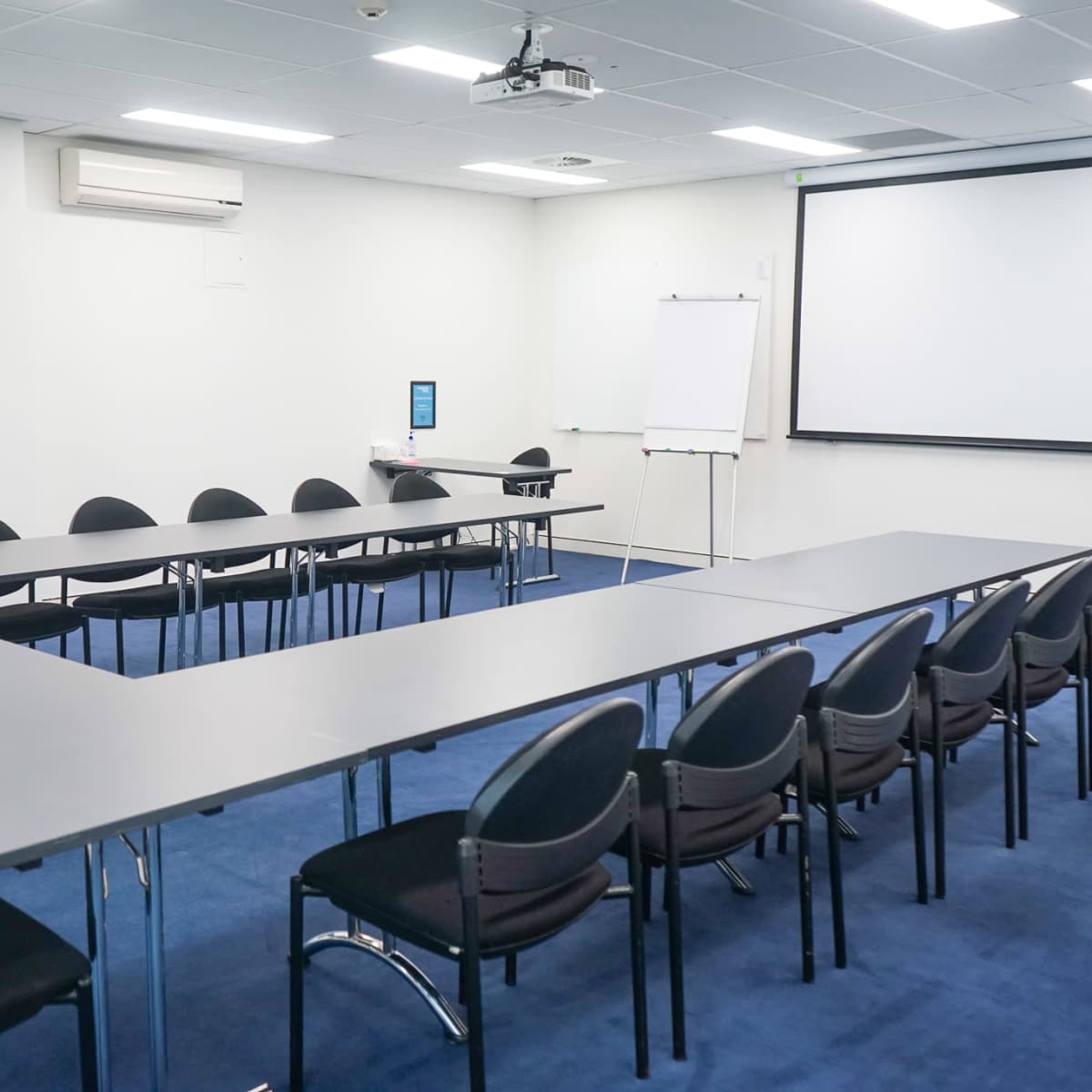 A conference room features a U-shaped arrangement of dark tables and black chairs, with a projector screen and whiteboard at one end.